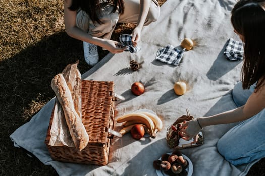 Two beautiful Caucasian girls are preparing to relax on a picnic with a basket of fruits, a French baguette and napkins on a blanket in the park on a summer day, flat lay close-up.