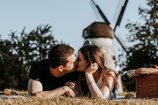 One beautiful Caucasian couple gently kisses each other on the lips on a sunny spring day while lying in the park on a picnic against the backdrop of an old mill, close-up view from below.