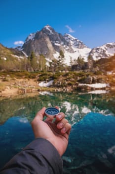 A man with a compass in his hand in high mountains near a clean lake. Travel concept. Landscape photography. Traveler man looking for direction with compass on coast near lake in mountains, first person view.