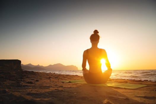 A woman meditates on the coast by the sea, rear view. Place for text. Yoga, health harmony, silhouette of a young woman on the beach at sunset or dawn.
