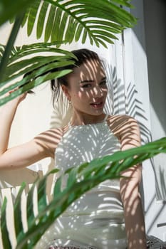 a brunette woman in light clothes sits by the door in the shade of a plant