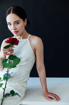 beautiful woman in a white dress sits with a red rose on a dark background