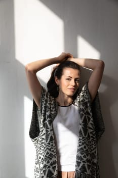 brunette woman standing in the shade of a window against a white wall posing