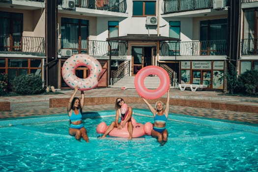 Three women are in a pool, holding pink inflatable donuts. Scene is fun and playful
