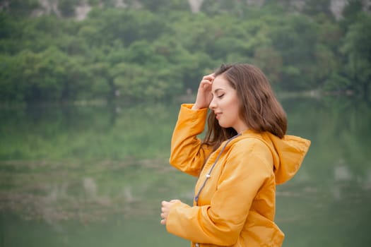 A woman in a yellow jacket is standing by a lake. She is looking at the water and she is lost in thought