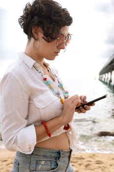 Vertical portrait woman on the beach with mobile phone in her hand typing smiling with sunglasses