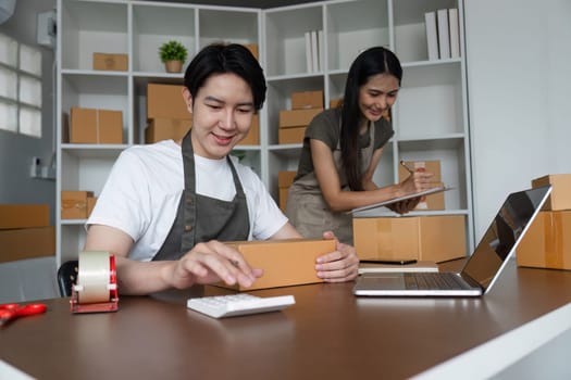 A couple working together in their home office, taking orders and packing products for online sales. Surrounded by cardboard boxes and using a laptop.