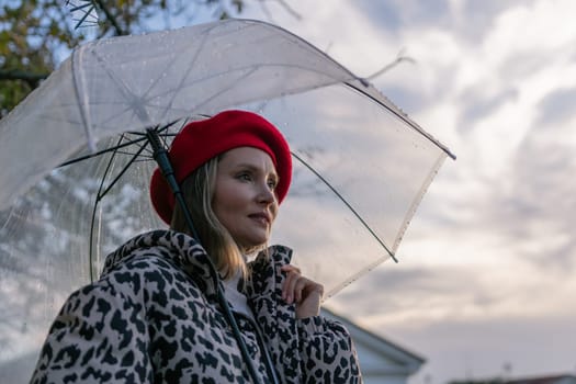 A woman wearing a red hat and a leopard print coat is holding a clear umbrella. The umbrella is open, and the woman is looking up at the sky. The scene has a calm and peaceful mood