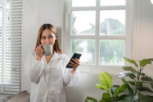 A young woman in a white shirt enjoys her morning routine, sipping coffee and using her smartphone by a window in a bright, modern home.