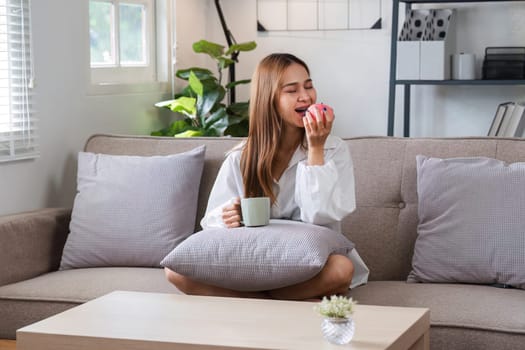 A young woman starts her day with a healthy morning routine, enjoying coffee and fruit while sitting on a cozy sofa in a modern living room.