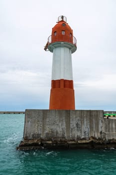 A white and orange lighthouse stands tall on a concrete pier in coastal waters. The lighthouse has a red roof and a metal railing. The water is a vibrant turquoise, and the sky is a light gray.