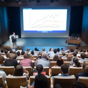 Business and entrepreneurship symposium. Speaker giving a talk at business meeting. Audience in conference hall. Rear view of unrecognized participant in audience.