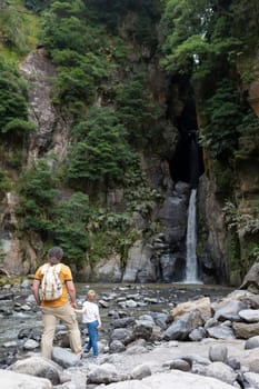A man and a child are standing near a waterfall. The man is wearing a yellow shirt and a backpack. The scene is peaceful and serene