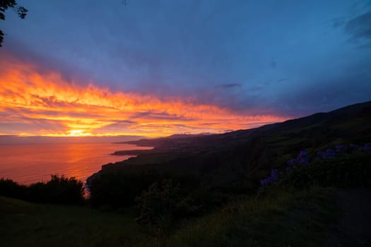 A beautiful sunset over the ocean with a mountain in the background. The sky is filled with clouds and the sun is setting