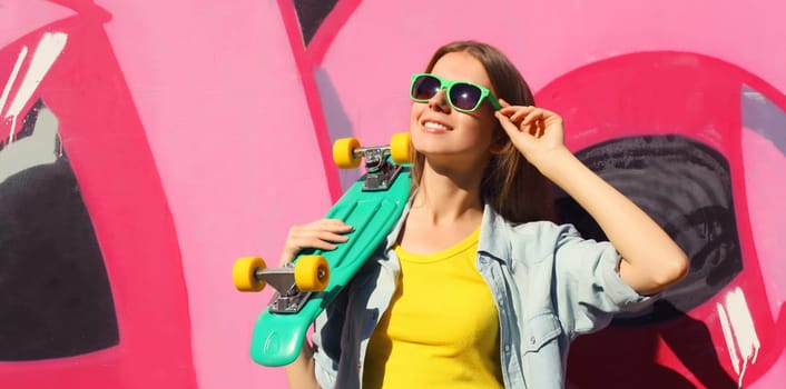 Stylish happy young girl with skateboard in glasses, modern teenager smiling girl looking away posing against the background of pink graffiti wall