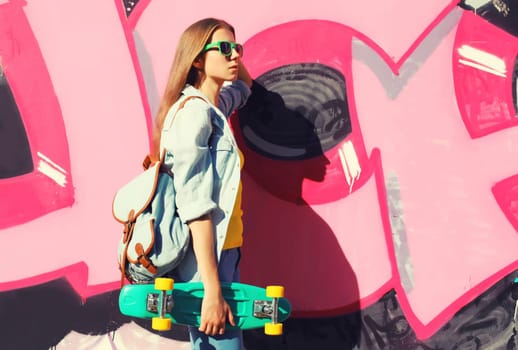 Stylish young girl with skateboard in glasses, modern teenager girl looking away posing against the background of pink graffiti wall