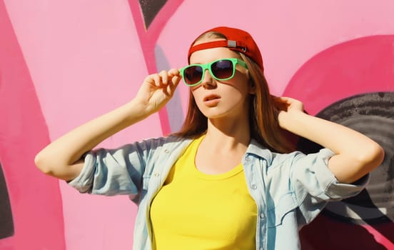 Portrait of stylish young girl in red baseball cap, glasses, modern teenager girl posing against the background of pink graffiti wall