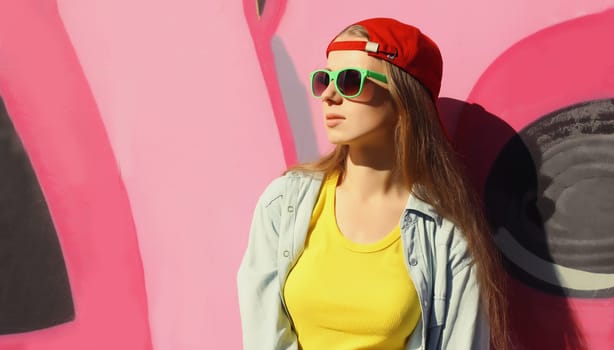 Portrait of stylish young girl in red baseball cap, glasses, modern teenager girl looking away posing against the background of pink graffiti wall