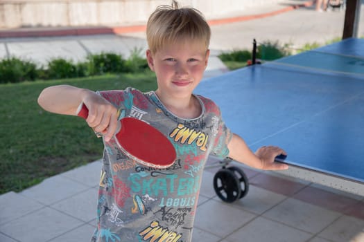 eight year old blond boy plays ping pong with a red racket on a special table tennis court, fun summer sports activities for kids,unforgettable summer holidays in Turkey