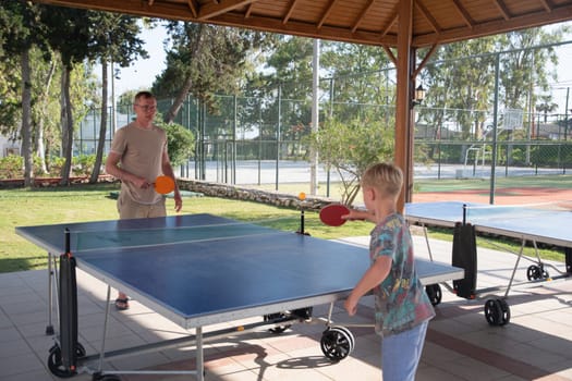 Young father plays teaching his 8 year old son to play ping pong on a hotel playground in Turkey, table tennis, fun summer sports activities for kids