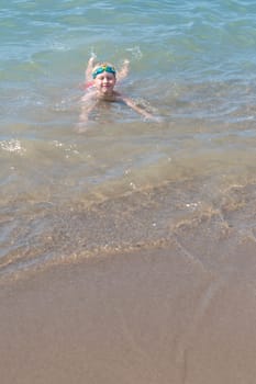boy 8 years old in swimming goggles lies in the surf on the sandy beach of the blue sea, loves to swim during the summer holidays in Turkey, refreshing swim on a hot sunny day