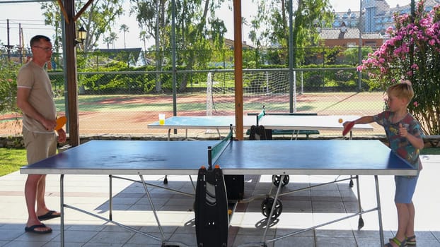 eight year old blond boy plays ping pong with his father on a special table tennis court, fun summer sports activities for children, unforgettable summer holidays with the whole family in Turkey