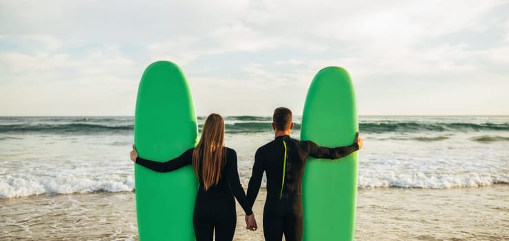 A couple in wetsuits stands on a sandy beach holding their green surfboards as the sun sets. They are holding hands and looking out towards the ocean, which is calm and has gentle waves, panorama