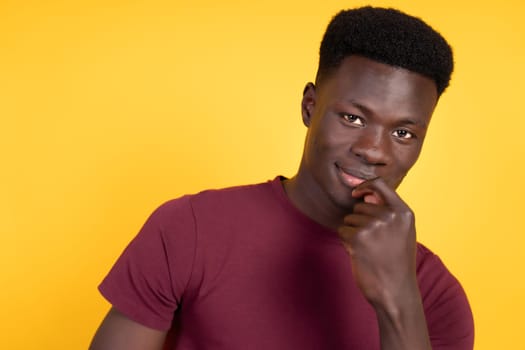 A young man with short black hair is seen against a vibrant yellow backdrop, wearing a red shirt and displaying a thoughtful expression. He has his hand resting gently on his chin, as if pondering deeply or considering a decision. With copy space.