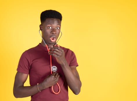 Attractive young black man wearing a red t-shirt and with a red stethoscope listening to his heart and looking surprised, with a yellow background and copy space.