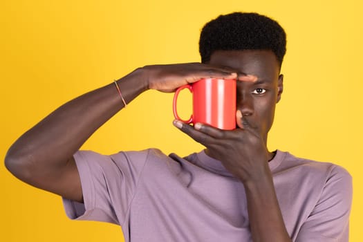 A man with a red mug in his hand is looking at the camera. The mug is placed in front of him, and he is holding it up to his face. Concept of curiosity and playfulness