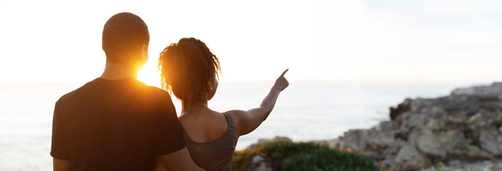 Young african american woman hugging man in sportswear, enjoy workout break, point finger at empty space in morning on rocks at ocean, outdoor. Relationship, health care, active lifestyle and sports