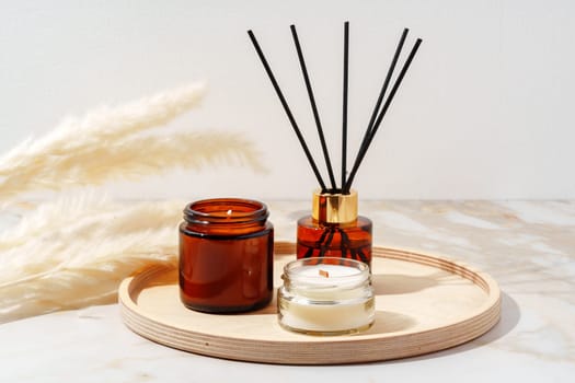 Three brown glass containers, one with black reeds and two with white candles, sit on a wooden tray on a marble surface.