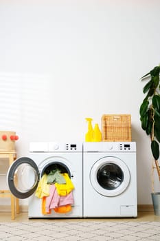 A modern laundry room with two white washing machines. One washing machine has the door open and colorful clothes hanging out of it.