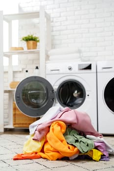 A pile of colorful, clean laundry sits on a patterned rug beside a white washing machine in a modern, light-colored laundry room.