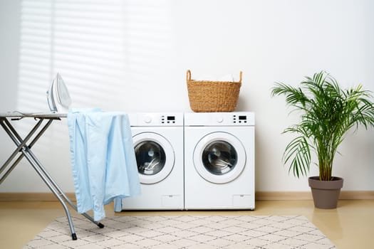 A modern laundry room with two white washing machines, a wicker basket containing laundry, and a potted plant in the corner.