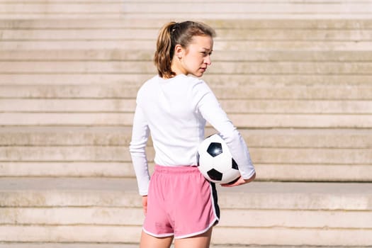 rear view of a sports woman in a concrete soccer court with a ball under her arm, concept of football lifestyle and urban sport in the city, copy space for text
