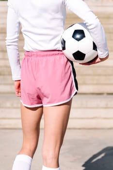 rear view of an unrecognizable sports woman in a concrete soccer court with a ball under her arm, concept of football lifestyle and urban sport in the city