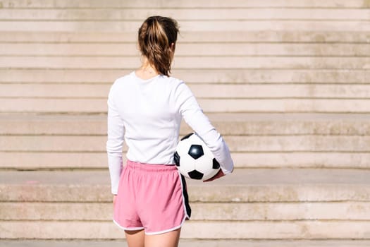 rear view of an unrecognizable sports woman in a concrete soccer court with a ball under her arm, concept of football lifestyle and urban sport in the city, copy space for text