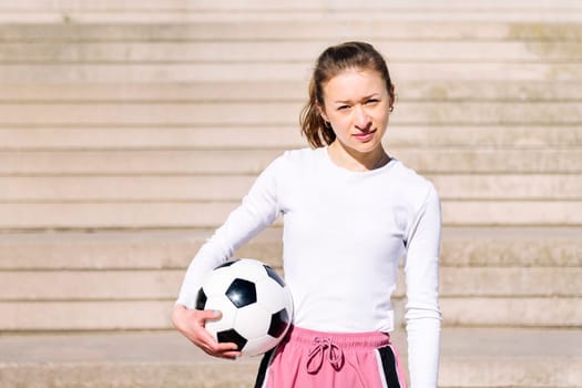 sports woman looking at camera in a concrete soccer court with a ball under her arm, concept of football lifestyle and urban sport in the city, copy space for text