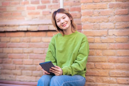 Beautiful Chinese teen student smiling using social media app on digital tablet looking at camera outdoors. Girl sitting chatting on digital tablet