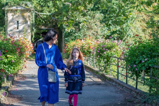 young mother escorts her little daughter to school,together they walk through the park,the student is dressed in blue uniform jacket with skirt and pink tights, back to school on a sunny day