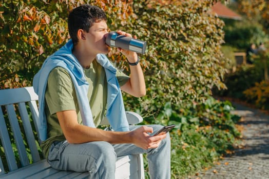 a teenager sits on a bench in the park drinks coffee from a thermo mug and looks into a phone. Portrait of handsome cheerful guy sitting on bench fresh air using device browsing media smm drinking latte urban outside outdoor.