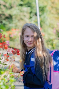 Back to school, portrait of a little girl from primary school outdoors, enjoying flowers in a flowerbed and looking at the camera with a smile, student dressed in dark blue uniform