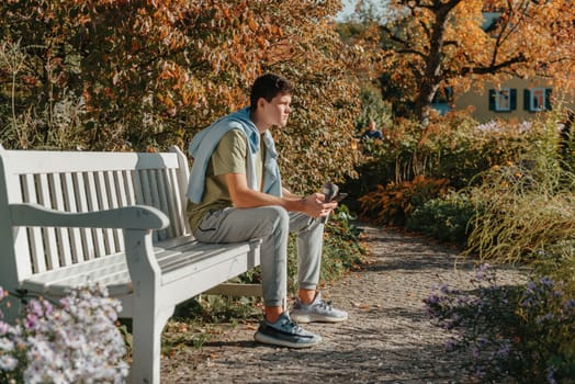 a teenager sits on a bench in the park drinks coffee from a thermo mug and looks into a phone. Portrait of handsome cheerful guy sitting on bench fresh air using device browsing media smm drinking latte urban outside outdoor.