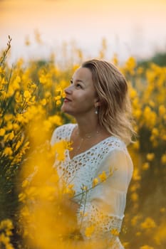 A woman is standing in a field of yellow flowers. She is wearing a white dress and a necklace. The flowers are in full bloom, creating a bright and cheerful atmosphere