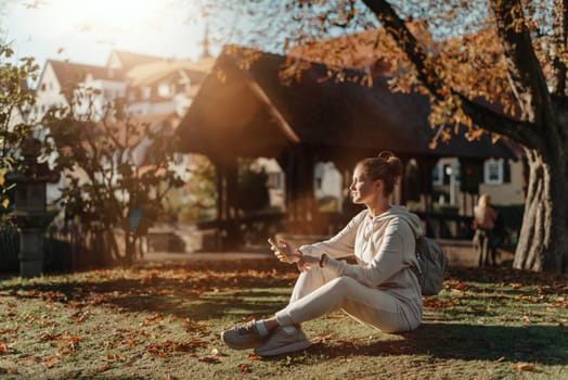 Young fashionable teenage girl with smartphone in park in autumn sitting at smiling. Trendy young woman in fall in park texting. Retouched, vibrant colors. Beautiful blonde teenage girl wearing casual modern autumn outfit sitting in park in autumn. Retouched, vibrant colors, brownish tones.