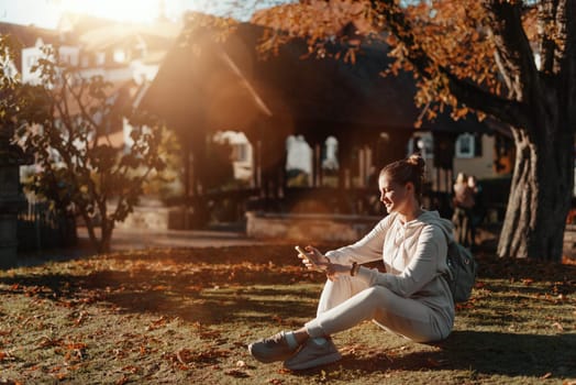 Young fashionable teenage girl with smartphone in park in autumn sitting at smiling. Trendy young woman in fall in park texting. Retouched, vibrant colors. Beautiful blonde teenage girl wearing casual modern autumn outfit sitting in park in autumn. Retouched, vibrant colors, brownish tones.