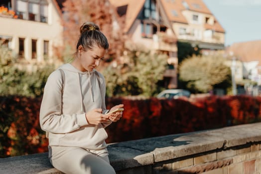 Young fashionable teenage girl with smartphone in park in autumn sitting at smiling. Trendy young woman in fall in park texting. Retouched, vibrant colors. Beautiful blonde teenage girl wearing casual modern autumn outfit sitting in park in autumn. Retouched, vibrant colors, brownish tones.