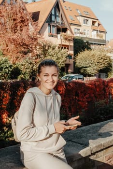 Young fashionable teenage girl with smartphone in park in autumn sitting at smiling. Trendy young woman in fall in park texting. Retouched, vibrant colors. Beautiful blonde teenage girl wearing casual modern autumn outfit sitting in park in autumn. Retouched, vibrant colors, brownish tones.