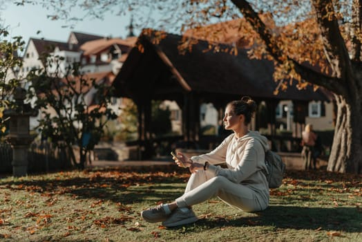 Young fashionable teenage girl with smartphone in park in autumn sitting at smiling. Trendy young woman in fall in park texting. Retouched, vibrant colors. Beautiful blonde teenage girl wearing casual modern autumn outfit sitting in park in autumn. Retouched, vibrant colors, brownish tones.